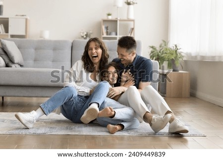 Cheerful excited parents tickling beloved kid girl, playing with child on heating floor in modern home interior, sitting on carpet, laughing, shouting for joy, enjoying family leisure Royalty-Free Stock Photo #2409449839
