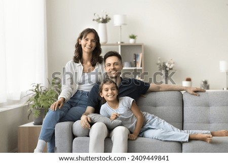Cheerful young parents and cute tween girl kid relaxing together on soft couch at cozy home, looking at camera with toothy smiles, enjoying leisure in stylish new apartment. Family portrait