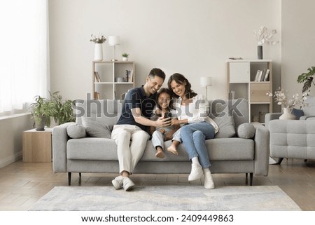 Positive young mom, dad and sweet girl kid taking family selfie on cozy comfortable couch, having fun with digital gadget in cozy stylish home interior, using smartphone for selfie together