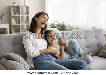 Happy excited young mom and cheerful daughter kid having fun on soft couch at home, relaxing on sofa, looking away, laughing, enjoying family leisure, comfort, relaxation, close relationships Royalty-Free Stock Photo #2409449869