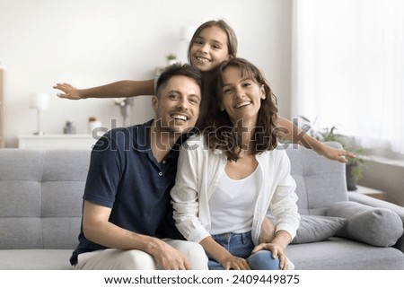 Happy loving parents sitting on home couch close together. Cheerful kid girl leaning on mom and dad from behind, playing airplane with open arms, looking at camera for family portrait, smiling