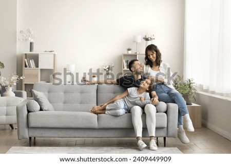 Happy couple of young parents and little daughter kid sitting and relaxing on sofa, resting on comfortable couch in modern cozy home interior, talking, laughing, smiling. Full length wide shot