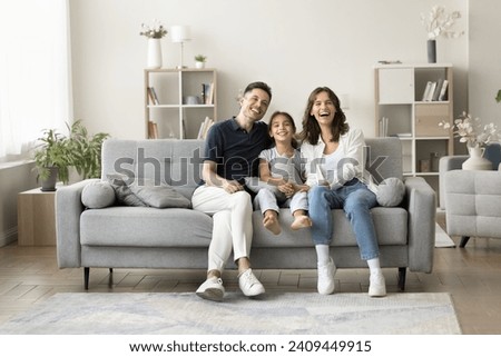 Joyful young parents and sweet kid girl sitting on soft comfortable grey couch in cozy home interior, looking at camera, laughing, smiling, promoting mortgage, property buying. Full length, wide shot