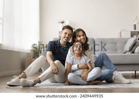 Cheerful family couple holding beautiful tween daughter girl in arms, sitting on heating floor in modern home interior, looking at camera for portrait, smiling, enjoying leisure in new apartment Royalty-Free Stock Photo #2409449935