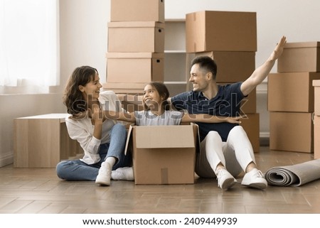Playful beautiful girl kid playing airplane in moving box, sitting in paper packages with open arms. Cheerful mom, dad and child celebrating relocation at heap of stacked containers Royalty-Free Stock Photo #2409449939
