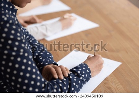 Close up of diverse concentrated student sit at desk in classroom writing examination or test, focused multiethnic group busy summarizing lecture or lesson, handwriting making notes on paper Royalty-Free Stock Photo #2409450109