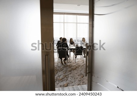 Diverse young business team sitting at large table in meeting room, talking, discussing collaboration on project, brainstorming on creative ideas. Full length candid shot, door view