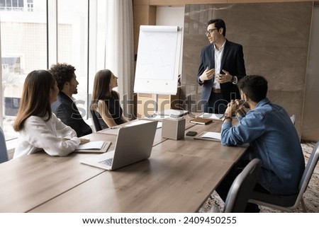 Serious young business leader man speaking to diverse team of colleagues, standing at meeting table, telling project plan, management strategy, ideas for brainstorming