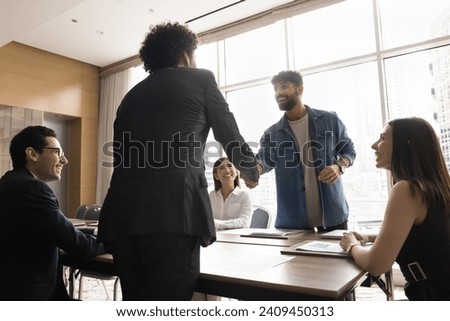 Cheerful young Arab startup leader and investor getting agreement on partnership meeting, shaking hands over table, smiling, laughing, discussing successful business cooperation Royalty-Free Stock Photo #2409450313