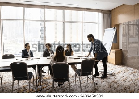 Young Arab project manager man presenting startup to colleagues, standing at large meeting table at leader place. Coach giving seminar, lecture in modern office boardroom interior Royalty-Free Stock Photo #2409450331