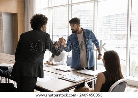 Two office competitors men fighting in arm wrestling battle over table, arguing at diverse team meeting, expressing disagreement, having problems with teamwork, competing for leadership