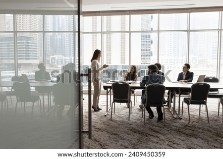 Wide shot of multiethnic business team talking at meeting table in office conference room, brainstorming in co-working space with large window. Project leader woman talking to colleagues Royalty-Free Stock Photo #2409450359