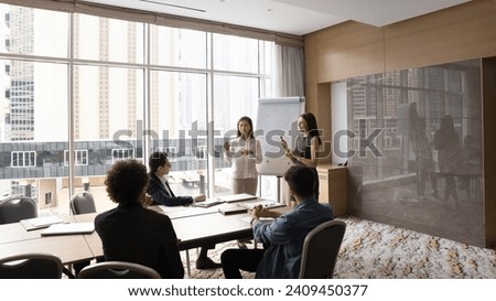 Two diverse speaker women presenting marketing report on whiteboard to coworkers, managers, speaking at flipchart, giving training, presentation in modern office meeting room interior Royalty-Free Stock Photo #2409450377