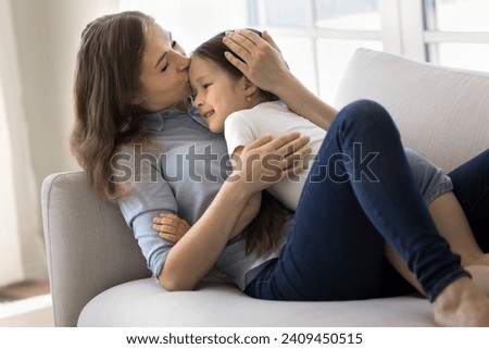 Loving mother express care, showing support, feeling unconditional love lying on sofa with adorable daughter, kissing small kid girl forehead relaxing together on weekend at home. Motherhood, cherish