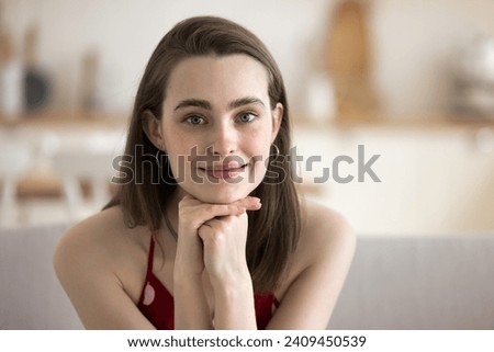 Head shot portrait of cute young woman staring at camera, seated on couch in studio flat spend time alone at own or rented apartment, having attractive appearance. Younger generation female portrait