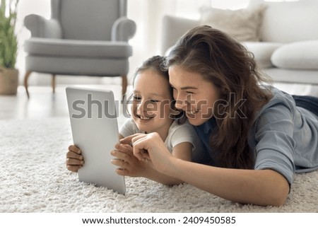 Happy young mother spend time with little daughter at home, lie on floor at home have fun use digital tablet together, watch cartoons, funny video content in social networks, enjoy modern application