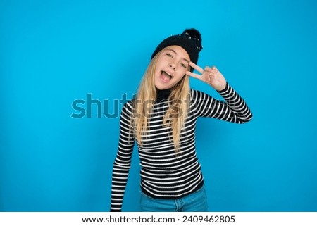 beautiful caucasian teen girl knitted  hat and striped T-shirt  Doing peace symbol with fingers over face, smiling cheerful showing victory