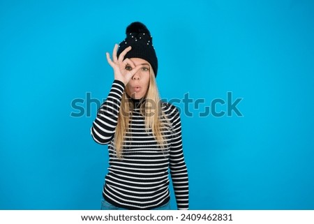 beautiful caucasian teen girl knitted  hat and striped T-shirt doing ok gesture shocked with surprised face, eye looking through fingers. Unbelieving expression.