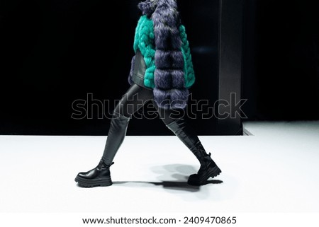  Female figure dressed in a fur coat and black leather boots walking on black background.
