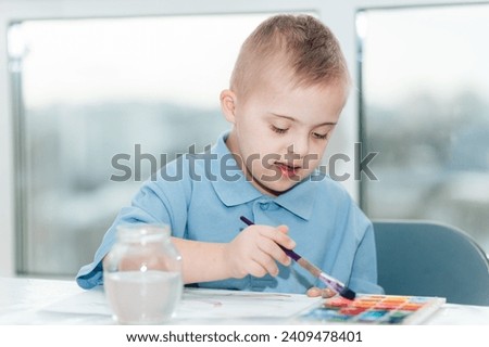 A boy with Down syndrome draws with watercolors in a school album. The child is creative. Early development and preparation for school.
