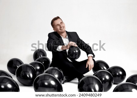 elegant caucasian man in suit tuxedo sitting on studio floor with a lot of black air balloons Royalty-Free Stock Photo #2409480249
