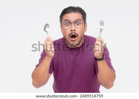 A bespectacled middle aged man looking awestruck while looking at something mouthwatering. Holding a fork and spoon hungry for a feast. Isolated on a white background. Royalty-Free Stock Photo #2409514759