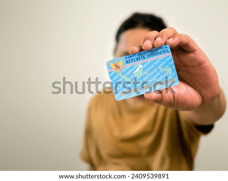 A person shows and holds an identity card (KTP) wearing a brown T-shirt and a plain background. Selective focus