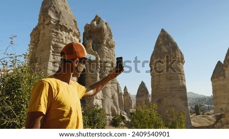 A man in a yellow T-shirt and baseball cap takes a picture of the mountains with his phone. Love Valley in Cappadocia. A blogger shoots a story on a smartphone about cliffs in Turkey