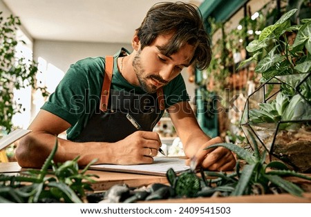 Inventory at floristic shop. Focused caucasian man leaning on desk and writing on clipboard while working at modern store. Bearded florist running successful nursery with green domestic plant.