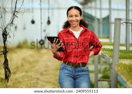 Multi-ethnic female farmer standing in a greenhouse, holding tablet, smiling.