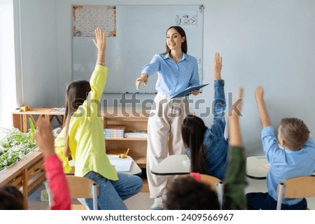 Friendly female teacher in classroom teaches elementary school students, kids raising hands to teacher aasks questions to small students sitting at their desks Royalty-Free Stock Photo #2409566967