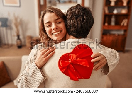 Happy Valentine's Day. A man gives a heart-shaped gift box to his beloved woman in the living room at home, the woman hugging him tenderly. Romantic evening together. Royalty-Free Stock Photo #2409568089