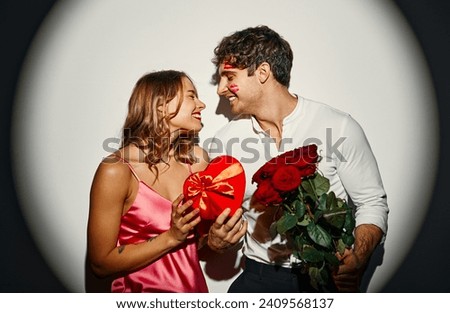 Happy Valentine's Day. Couple in love standing with a heart-shaped gift box and a bouquet of roses on a white background under the light of a spotlight.