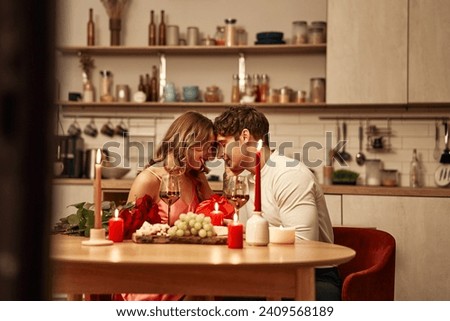 Happy Valentine's Day. A couple with glasses of wine by candlelight sitting in the kitchen at the table, romantically spending the evening together. A man giving his beloved a heart-shaped gift box. Royalty-Free Stock Photo #2409568189