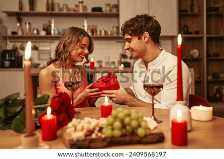 Happy Valentine's Day. A couple with glasses of wine by candlelight sitting in the kitchen at the table, romantically spending the evening together. A man giving his beloved a heart-shaped gift box. Royalty-Free Stock Photo #2409568197