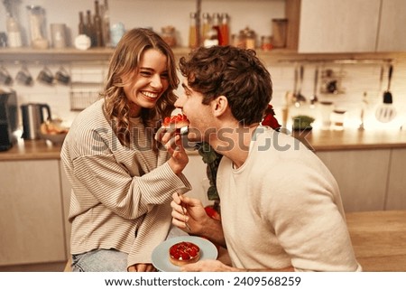 Happy Valentine's Day. Young couple in love eating cakes while sitting on the table in the kitchen, romantically spending the evening together. Royalty-Free Stock Photo #2409568259