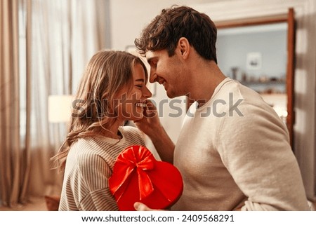 Happy Valentine's Day. A man gives a heart-shaped gift box to his beloved woman in the living room at home, the woman hugging him tenderly. Romantic evening together.