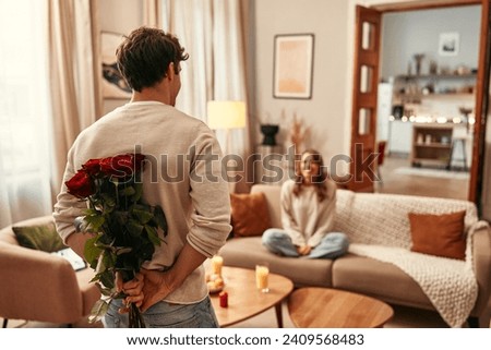 Happy Valentine's Day. A man holding a bouquet of red roses behind his back for his beloved woman who is sitting on the sofa in the living room at home. Royalty-Free Stock Photo #2409568483