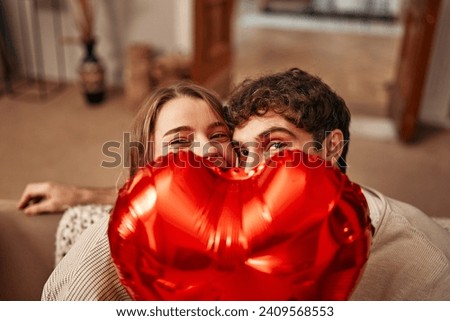 Happy Valentine's Day. Young couple in love holding a heart-shaped balloon, hiding behind it while sitting on the sofa in the living room at home. Romantic evening together. Royalty-Free Stock Photo #2409568553