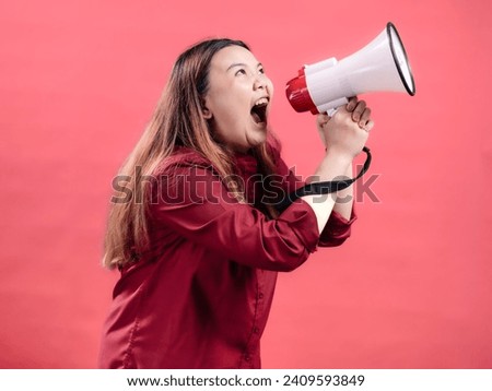 Portrait of an Indonesian Asian woman wearing a red dress, speaking loudly through a megaphone, isolated against a pink background.