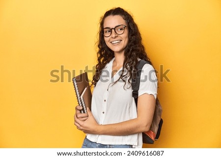 Student holding notebooks, glasses, backpack on, looks aside smiling, cheerful and pleasant. Royalty-Free Stock Photo #2409616087