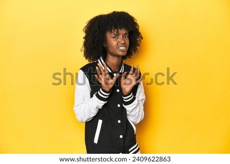 Teen girl in baseball jacket, yellow studio background rejecting someone showing a gesture of disgust.