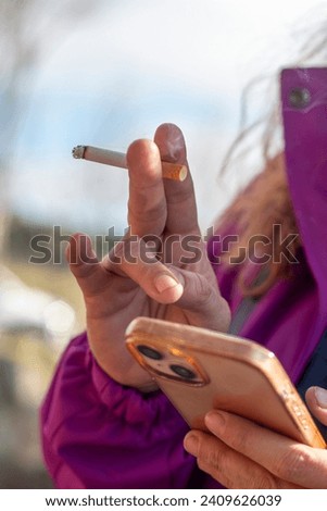 Woman's hand holding a smoking cigarette while using her mobile phone. Concept: vice Royalty-Free Stock Photo #2409626039