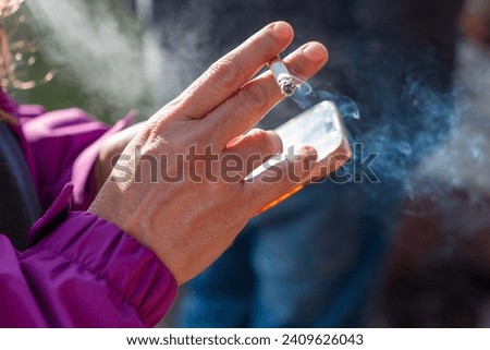 Woman's hand holding a smoking cigarette while using her mobile phone. Concept: vice Royalty-Free Stock Photo #2409626043
