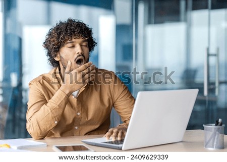 Young overworked and overtired businessman yawning, working inside office with laptop, boring long routine work, man in casual clothes. Royalty-Free Stock Photo #2409629339