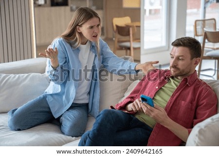 Indignant, offended, hysterical woman screams at husband sitting on sofa with cellphone. Couple family quarrel conflict misunderstanding discord. Indifferent man ignores dissatisfied wife's questions. Royalty-Free Stock Photo #2409642165