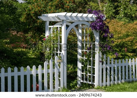 A white wooden archway and white wood picket fence surround a garden. There's a colorful purple vine hanging along the top of the fence and around the arch. The garden has trees and lush green grass. Royalty-Free Stock Photo #2409674243