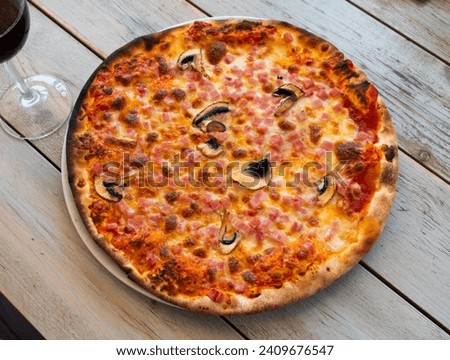 On plate is whole round freshly baked crispy pizza with mushrooms champignon and cubes of pork ham.Dish is complemented with glass of dry red wine