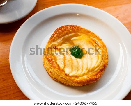 On plate is round little dessert - apple pie made of puff pastry with slices of natural apples,decorated with green candied fruit Royalty-Free Stock Photo #2409677455