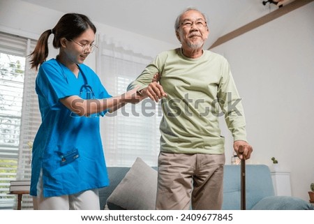 International Day for the Elderly. Young carer supporting helping senior man stand up with walking stick from sofa, Nurse wearing blue uniform help helping his patient senior old man with walking cane